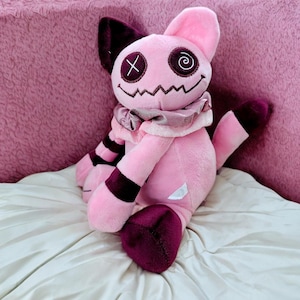 May include: A pink plush toy with a cat-like appearance. It has dark purple accents on the ears, paws, and tail. The toy features stitched details, including an X for one eye and a spiral for the other. The mouth is a zig-zag shape, and it has a ruffled collar.