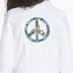 May include: White denim jacket with a peace sign patch on the back. The peace sign is made of denim patches in various shades of blue, with colorful floral accents in the center. The jacket is worn with light blue jeans.