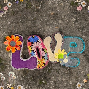 May include: A colorful, fabric-based word art piece spelling out "LOVE". The letters are adorned with floral embellishments and a variety of textures, including denim and felt. The background is a textured gray surface, with floral accents.