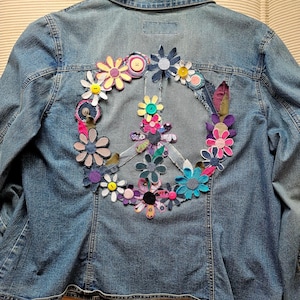 May include: A light blue denim jacket with a peace sign made of colorful fabric flowers and buttons. The flowers are in shades of pink, yellow, blue, and purple. The jacket has a classic collar and button closure.