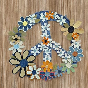 May include: A denim peace sign decorated with various fabric flowers in shades of white, orange, blue, and green. The peace sign is set against a wooden background.