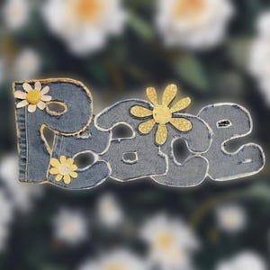 May include: A denim-textured sign spelling out the word "peace" in a whimsical font. The letters are embellished with yellow and white daisy-like flowers. The sign is set against a blurred floral background.