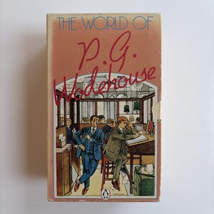 May include: A rectangular book box set titled The World of P.G. Wodehouse. The cover features a colorful illustration of men in suits working in an office setting, with a small penguin logo at the bottom center of the box.