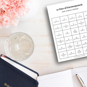 May include: A still-life arrangement featuring a navy blue Holy Bible, a clear glass, a notebook, and a pen. A calendar titled "30 Days of Encouragement" is visible. Pink flowers in a white vase add a soft touch.