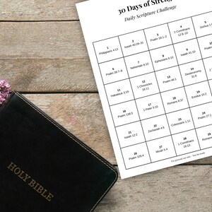 Könnte beinhalten: Eine schwarze, ledergebundene Bibel liegt auf einer Holzoberfläche neben einem weißen Blatt mit dem Text "30 Days of Strength Daily Scripture Challenge". Das Blatt zeigt ein Raster mit Bibelversen.