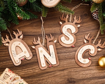 May include: Wooden Christmas ornaments shaped like reindeer heads, each with a letter or name cutout. The letters are A, N, S, and C, with names Astrid, Norton, Simon, and Cora. The ornaments have snowflake details and red noses.