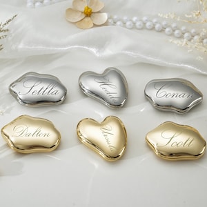 May include: Six polished metal stones in silver and gold tones, each engraved with a unique name: Settla, Hedda, Conan, Dalton, Ursula, and Scott. These smooth, irregular heart-shaped objects feature a reflective finish and elegant cursive lettering.