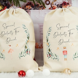 Puede incluir: Dos bolsas de regalo crema con cordón con el texto "Special Delivery For David". Una bolsa presenta una bailarina, la otra un cascanueces. Ambas bolsas tienen un diseño de corona verde y floral.