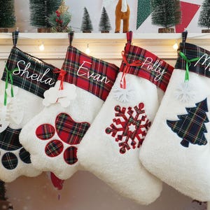 Puede incluir: Cuatro calcetines navideños blancos con puños a cuadros rojos y verdes, personalizados con los nombres Sheila, Evan, Polly y Miles. Cada calcetín presenta un diseño único, incluyendo huellas de patas, un copo de nieve y un árbol de Navidad.