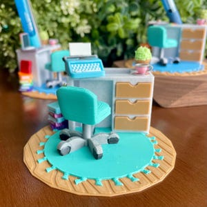 Puede incluir: Escena de escritorio en miniatura con una silla turquesa, una máquina de escribir y un escritorio con cajones. El escritorio está sobre una alfombra redonda con un centro turquesa y un borde beige. También se ve una pequeña pila de libros y un bolígrafo.