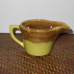 May include: A small, ceramic creamer with a handle and a spout. The creamer is two-toned, with a brown top and a yellow-green base. The handle is a matching yellow-green color.