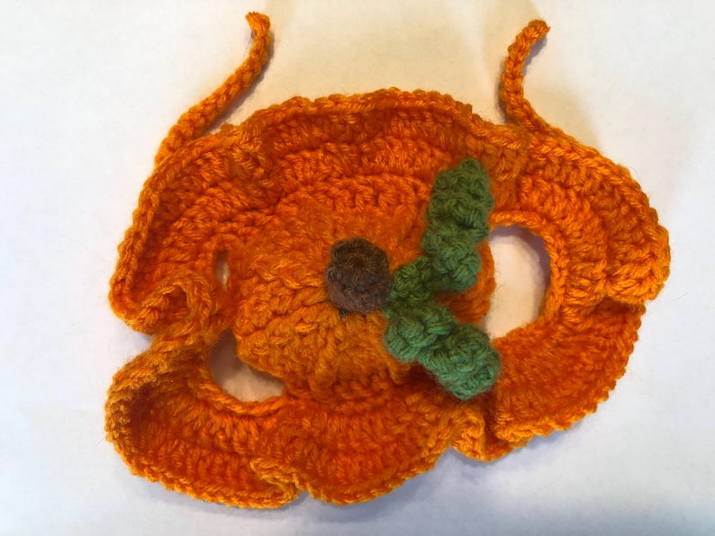 May include: A bright orange crocheted pumpkin accessory featuring a ruffled edge, a small brown centre stem, and a textured green leaf attached to the top. The piece includes two thin crocheted ties extending from the upper sides.