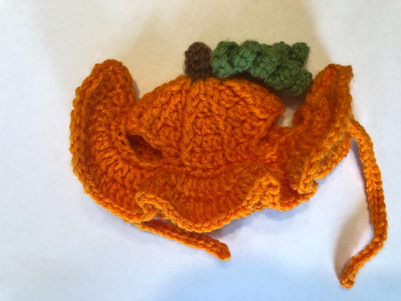 May include: A bright orange crocheted hat featuring a ruffled brim and a small brown stem with a textured green leaf attached to the top. The hat includes two long orange crocheted ties extending from the sides for securing the item in place.