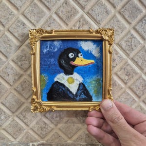 May include: A small, square painting of a duck in a gold ornate frame. The duck has a black head, yellow beak, and is wearing a white collar with a gold medallion. The background is blue with white clouds and green foliage.