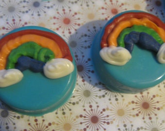 May include: Two blue and turquoise round cookies decorated with a rainbow design. The rainbow is made of orange, green, and blue frosting. The cookies are topped with white frosting clouds.