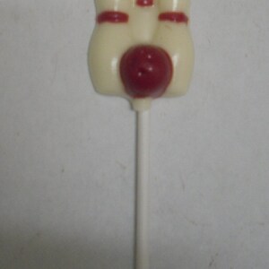 May include: White chocolate lollipop shaped like three bowling pins with a red ball in the center.