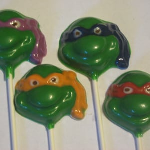 May include: Four green chocolate lollipops shaped like Teenage Mutant Ninja Turtle heads. Each lollipop has a different colored mask: purple, blue, orange, and red.