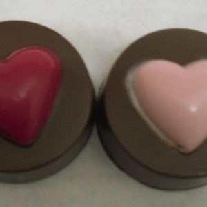May include: Two chocolate covered candies, each with a heart-shaped design. One heart is pink and the other is red.