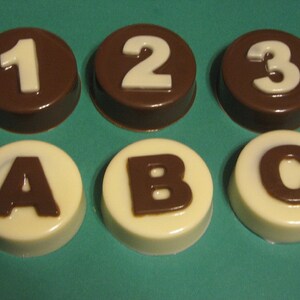May include: Six chocolate candies with white numbers and letters. The top row features the numbers 1, 2, and 3 on a dark chocolate base. The bottom row has the letters A, B, and C on a white chocolate base.