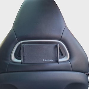 Puede incluir: Asiento de coche negro con marco de metal plateado y soporte para teléfono negro. El soporte del teléfono tiene el logotipo "Dunlop" impreso. El asiento es de un material oscuro y liso.