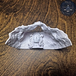 K&ouml;nnte beinhalten: Eine hellgraue Skulptur eines Medusenhauptes, umrahmt von einem felsigen Torbogen. Die Skulptur weist detaillierte Gesichtsz&uuml;ge und schlangenartiges Haar auf. Das Produkt wird auf einer Holzoberfl&auml;che pr&auml;sentiert, mit dem Text "Broken Top Prints".