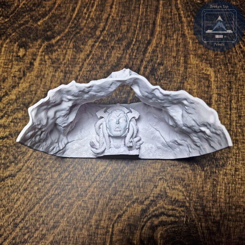 K&ouml;nnte beinhalten: Eine hellgraue Skulptur eines Medusenhauptes, umrahmt von einem felsigen Torbogen. Die Skulptur weist detaillierte Gesichtsz&uuml;ge und schlangenartiges Haar auf. Das Produkt wird auf einer Holzoberfl&auml;che pr&auml;sentiert, mit dem Text "Broken Top Prints".
