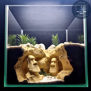 May include: A clear glass terrarium with a miniature landscape. The scene includes two beige Moai statues, artificial plants, and a rock formation. The terrarium has a black background and the text "Broken Top Prints" in the upper right corner.