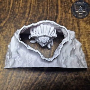 K&ouml;nnte beinhalten: Eine graue, 3D-gedruckte Skulptur einer Kreatur, die aus einer felsigen H&ouml;hle auftaucht. Die Kreatur hat einen detaillierten, insektenartigen Kopf und K&ouml;rper. Die H&ouml;hle ist strukturiert und hat ein raues, nat&uuml;rliches Aussehen. Das Logo "Broken Top Prints" befindet sich oben rechts.