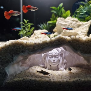 May include: An aquarium scene featuring several fish, including red-tailed guppies and neon tetras, swimming around a white Medusa sculpture. The sculpture is partially buried in sand, creating a unique underwater environment. The tank also includes plants and decorative rocks.