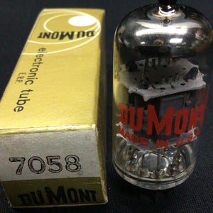 May include: A clear glass electronic vacuum tube with internal metal components and the red text DUMONT MADE IN U.S. printed on the side, positioned next to a gold cardboard box labeled DUMONT electronic tube E.R.P. 7058.