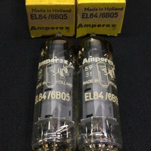 May include: Two Amperex EL84/6BQ5 electron tubes with clear glass envelopes and metal pins at the base. Each tube is paired with its original yellow cardboard box labelled Electron Tubes, Amperex, and Made in Holland with model EL84/6BQ5.