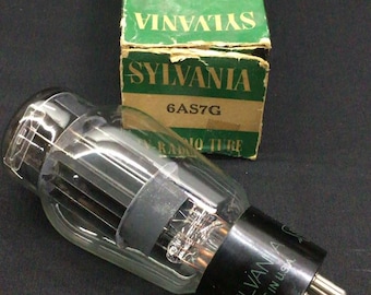 May include: A Sylvania 6AS7G vacuum tube with a clear glass bulb and a black base featuring metal pins. The tube is positioned in front of its original green and black cardboard box, which is labeled Television and Radio Tube Sylvania 6AS7G.