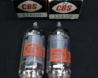 May include: Two clear glass vacuum tubes with metal pins at the base and orange labels, positioned in front of two matching black and red cardboard boxes. Each box features the text CBS and 12AU6 printed on the front and top panels.
