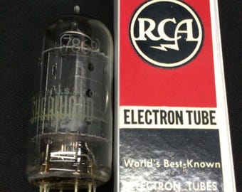 Może przedstawiać: Przezroczysta szklana lampa elektronowa RCA Radiotron z metalowymi pinami u podstawy, umieszczona obok prostokątnego białego, czerwonego i czarnego kartonowego pudełka. Pudełko zawiera logo RCA oraz tekst Radiotron, Electron Tube i World's Best-Known.