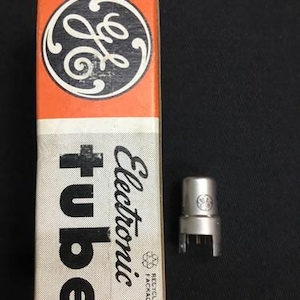 Puede incluir: Una caja naranja y blanca con el logotipo de GE y las palabras "tubo electrónico" impresas. Un tubo electrónico plateado está junto a la caja. La caja es rectangular y tiene un aspecto vintage.