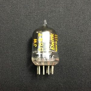 May include: A clear glass vacuum tube with the text "JW 5647 Electric MADE IN USA" in yellow. The tube has a rounded top and several pins at the base. The background is a dark gray fabric.