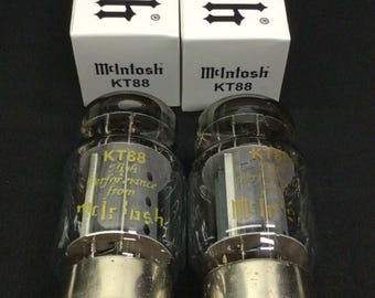 May include: Two McIntosh KT88 vacuum tubes with clear glass envelopes and metal bases, positioned in front of their matching white rectangular cardboard boxes. The boxes feature the McIntosh logo and KT88 text printed in black ink.