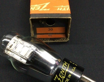 May include: A clear glass Zenith radio vacuum tube with a black base and metal pins, labeled Made in U.S.A. It is positioned in front of its original orange and black cardboard box, which features the Zenith brand name and the number 30 printed on it.