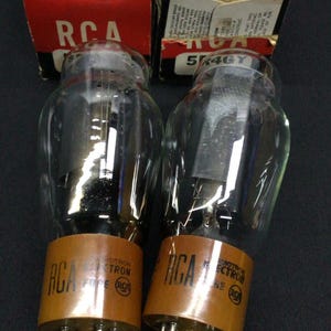 May include: Two RCA Radiotron 5K4GY vacuum tubes with clear glass bulbs and brown bases, each featuring metal pins at the bottom. The tubes are positioned in front of their original red and white cardboard boxes labeled with RCA branding.