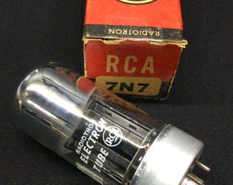 Może przedstawiać: Cylindryczna szklana lampa elektronowa RCA Radiotron 7N7 ze srebrną metalową podstawą i pinami, w komplecie z oryginalnym prostokątnym kartonowym pudełkiem. Pudełko jest czerwono-czarno-żółte, z logo RCA i napisem Electron Tube 7N7.