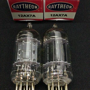Op de afbeelding: Twee Raytheon 12AX7A vacuümbuizen met heldere glazen omhulsels en interne metalen onderdelen, elk geplaatst voor hun originele blauwe, rode en witte kartonnen doos met het label Raytheon Television Tubes Sound and Sight.