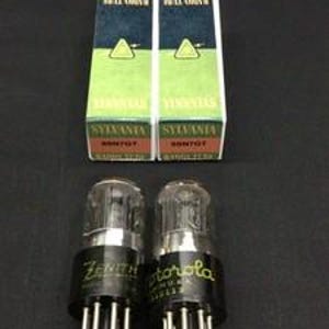 May include: Two vintage vacuum tubes with black bases and clear glass bulbs, labelled "Zenith" and "Motorola." Two boxes are behind the tubes, labelled "Radio Tube" and "Sylvania." The boxes have a green triangle design.