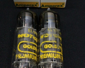 Może przedstawiać: Dwie przezroczyste szklane lampy próżniowe z metalowymi pinami u podstawy, każda z żółtymi etykietami z napisem EL84, 6BQ5, GOLD i PREMIUM. Znajdują się przed dwoma pasującymi białymi i żółtymi pudełkami z napisem Amperex Electron Tubes.