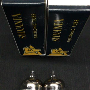 May include: Two Sylvania 5670 electronic tubes with their original boxes. The boxes are black with gold lettering and the Sylvania logo. The tubes are clear glass with silver internal components and multiple pins.