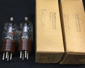 May include: Two clear glass vacuum tubes with brown bases and metal pins, positioned next to two rectangular tan cardboard boxes. The boxes are printed with the text Lewis Electronics, Acceptance Date, May 1945, U.S. Army-U.S. Navy, JAN-CN-3B24.
