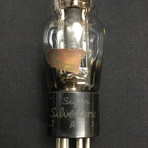 Peut inclure: Un tube à vide vintage avec une ampoule en verre transparent et une base noire. La base porte l'inscription "Super Silvertone .7 A X." Les composants internes sont visibles à l'intérieur du verre. Le tube a plusieurs broches argentées en bas.