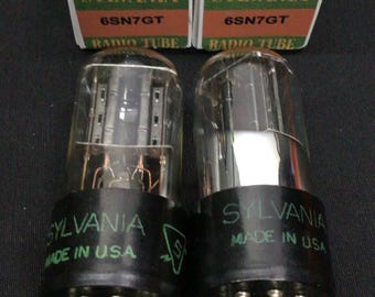 May include: Two Sylvania 6SN7GT radio tubes with clear glass envelopes and black bases, each labeled Sylvania Made in USA. They are accompanied by two matching green and white cardboard boxes featuring the Sylvania logo and text.