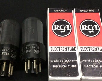 Może przedstawiać: Dwie szklane lampy elektronowe RCA Radiotron z czarnymi podstawami i metalowymi pinami, umieszczone obok dwóch pasujących prostokątnych biało-czerwonych kartonowych pudełek. Pudełka zawierają logo RCA oraz napis Radiotron Electron Tube.