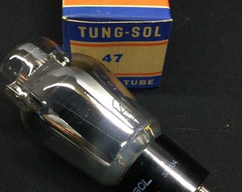 May include: A clear glass Tung-Sol radio tube with a black base and metal pins, positioned in front of its original rectangular cardboard box. The box is blue and orange with the text Tung-Sol Radio Tube and the number 47 printed on it.