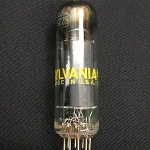 May include: A clear glass vacuum tube with the text "SYLVANIA MADE IN U.S.A." in yellow. The tube has a metallic top and multiple pins at the base. The background is a dark fabric.